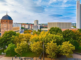 Grünfläche mit Bäumen und im Hintergrund mit hellblauem Himmel die Innenstadt von Chemnitz mit dem roten Turm